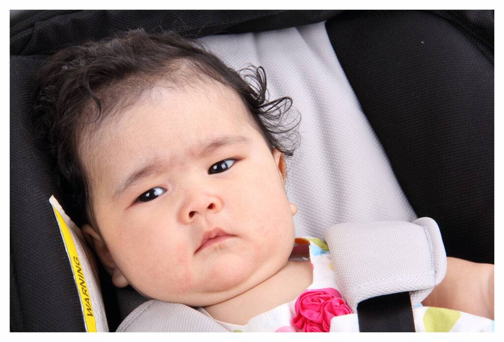Baby girl with mild eczema strapped into in car seat and looking grumpy
