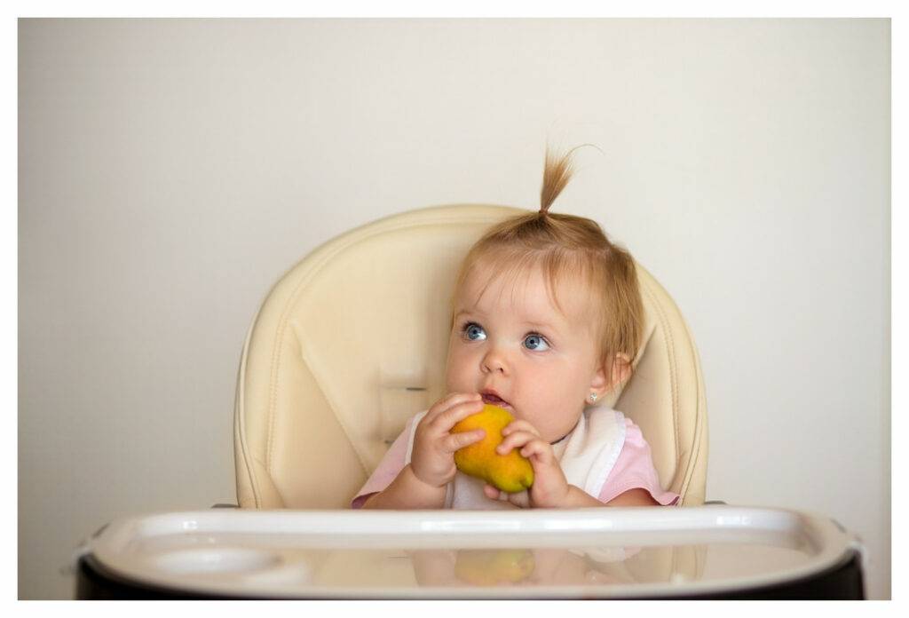 Little girl sat in high chair eating a pear. Pears are a good food for eczema as they contain vitamin K and vitamin C.