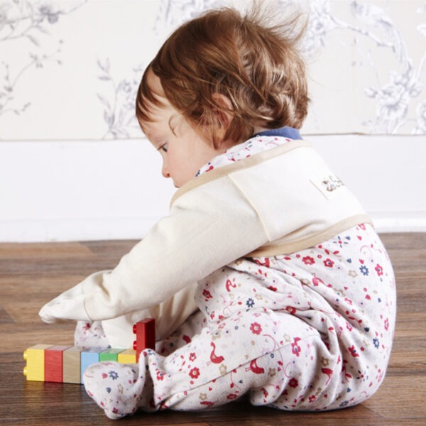 Side view of baby girl sat on floor playing with toys, wearing baby grow and babies bolero style original cream ScratchSleeves with cut away shoulders. Shows that the ScratchSleeves are a comfortable fit, not restrictive and can be worn over clothing.