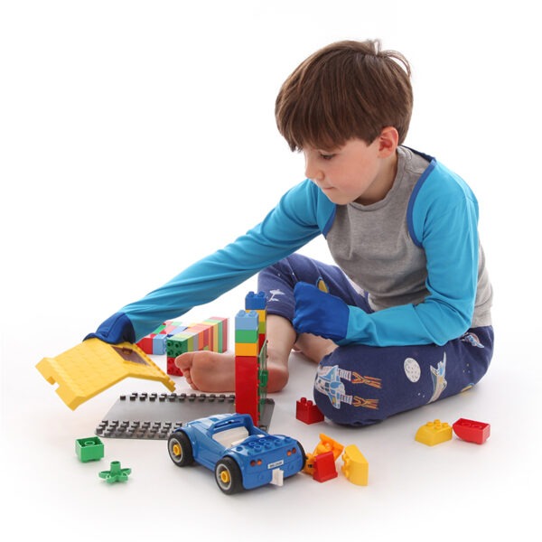 Young boy sat on floor playing with toys, wearing t-shirt, trousers and children's bolero style kingfisher blue special edition ScratchSleeves. Shows children can still play and use their hands naturally while their hands are covered by the silk eczema mitts. Also shows that the ScratchSleeves can be worn comfortably over clothing.