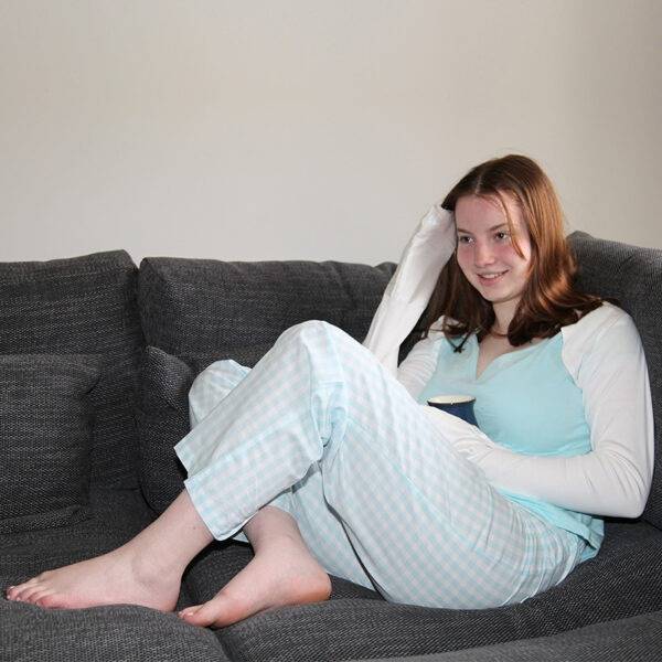 Young woman sat on sofa wearing pyjamas and white dye-free bolero style adults supersensitive ScratchSleeves, while holding a cup. Shows the ScratchSleeves are a relaxed, comfortable fit suitable for women and hands can still be used naturally even when covered by the silk eczema mitts.