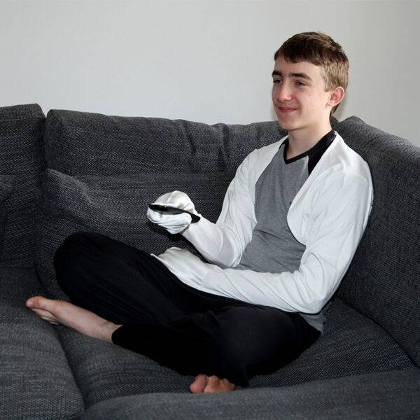Young man sat on sofa wearing pyjamas and white dye-free bolero style adults supersensitive ScratchSleeves, while holding remote control. Shows the ScratchSleeves are a relaxed, comfortable fit suitable for men and hands can still be used naturally even when covered by the silk eczema mitts.