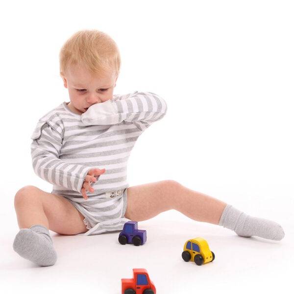 Toddler sat on floor playing with toys and wearing ScratchSleeves flip-mitt bodysuit. Eczema mitt is folded down to cover the left hand and the mitt on the right hand is folded back so the hand is free and the sleeve is hitched up with the GoodCatch tab. Shows the bodysuits are versatile and the mitts can be changed accordingly. The bodysuit has popper neck fastenings, white and grey marl striped body and sleeves with white mitts.