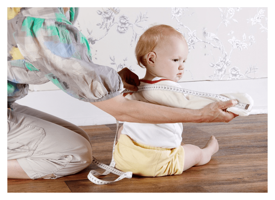 Mother measuring toddler's wingspan for correct sizing of ScratchSleeves. The child is facing away from the camera. The mother is holding the child's right hand stretching it away from the body, The tape measure is held under her thumb so that the end aligns with the child finger tips, the mothers other hand is stretching the tap measure to the centre of the child's back