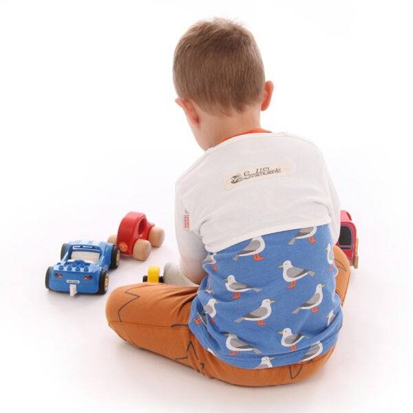 Back view of little boy sat on floor playing with toys, wearing t-shirt, trousers and children's bolero style white dye-free supersensitive ScratchSleeves. Shows that the ScratchSleeves are a comfortable fit, not restrictive and can be worn over clothing.