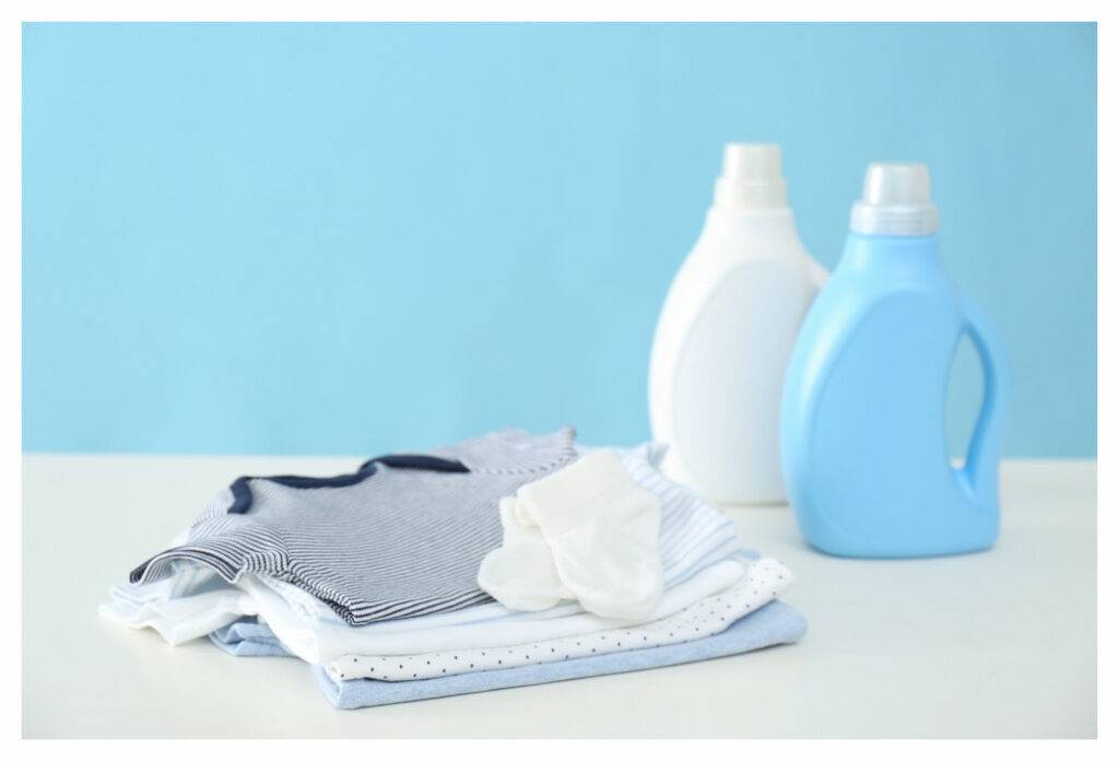 Pile of blue, grey and white folder baby clothes on counter with laundry detergent bottle in the background.
