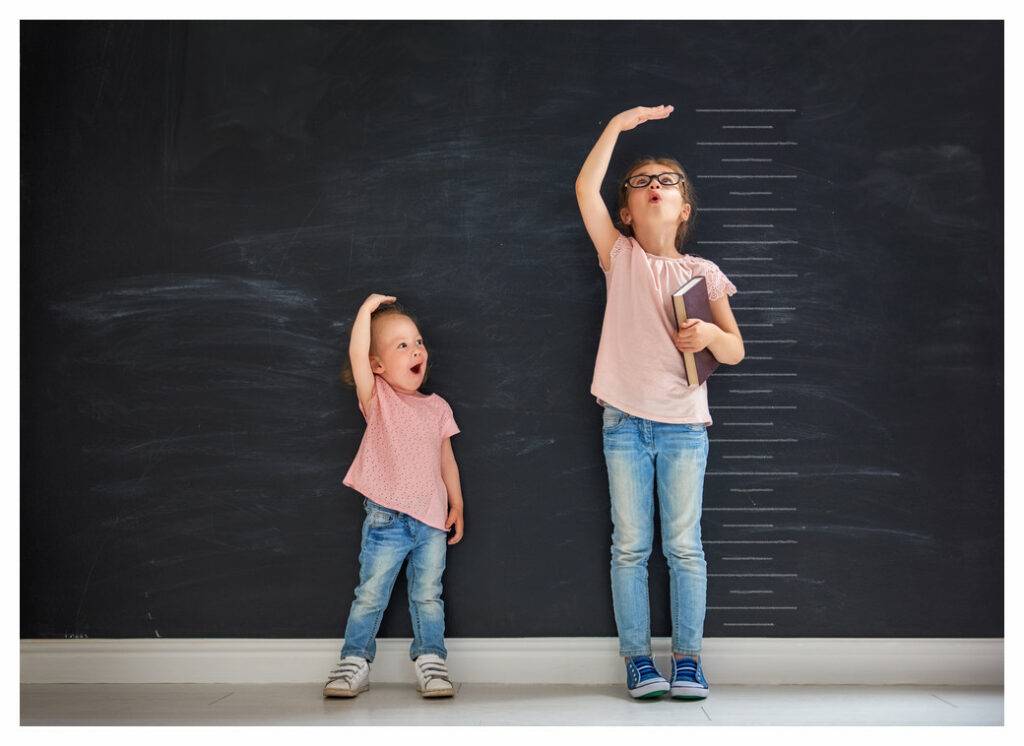 Young sisters in jeans and pink tops standing against a chalkboard with height chart. The sisters are comparing their relative heights. Both are over-estimating!
