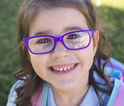 Little girl with funky purple glasses and a huge backpack. Her school uniform is crisp and new suggesting that she is just starting school.