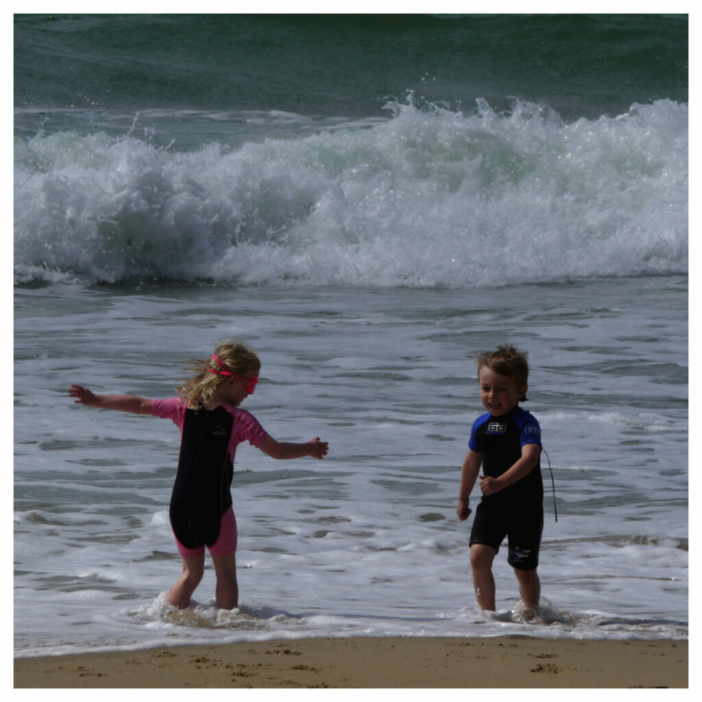 Two young children at the seaside, wearing wetsuits and playing the shallow water with waves breaking behind them.