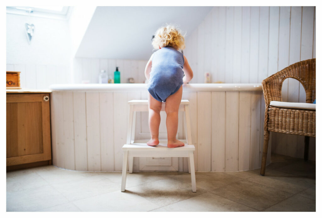 toddler wearing a blue onesie standing on steps (from IKEA) looking into a bathtub. There are a range of bath additives on the shelf behind the bath, but strangely no bath toys. 