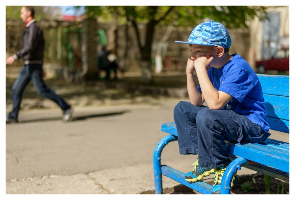 Bored tween boy sitting on park bench in the sun. He's resting his elbows on his knees and his chin on his hands.