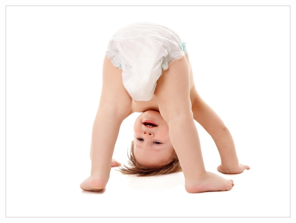 Toddler wearing a disposable nappy, standing on the floor and looking through their legs with hands and head also on the floor.