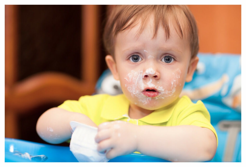 Toddler boy eating yoghurt. He's using his hands in the pot and his face is covered in yoghurt.