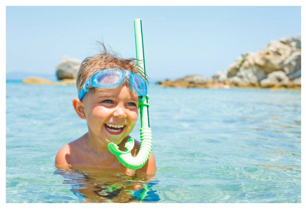 Small boy in the standing in clear sea water with a headland behind. He is wearing google and a snorkle