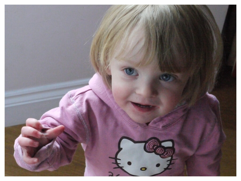 Toddler girl looking at the camera and baby signing 'milk'. She is making eye-contact and clearly communicating with the adult holding the camera.
