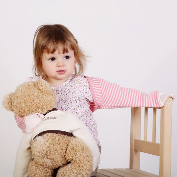 Little girl with teddy, holding onto chair and wearing top, trousers and pink stripe ScratchSleeves. Shows children can still play and use their hands naturally while their hands are covered by the silk eczema mitts.