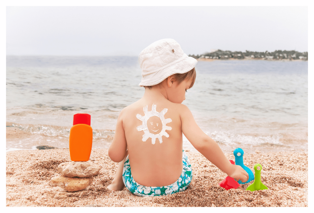 Young boy sat on sandy beach with a smiling sunshine painted on his back in white sunscreen. It looks like a typical English beach complete with overcast weather!