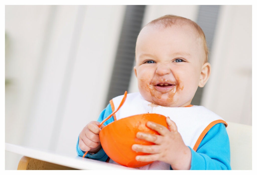 Young toddler sitting high chair holding spoon and bowl. wearing a bib and most of his lunch on his hands and face.