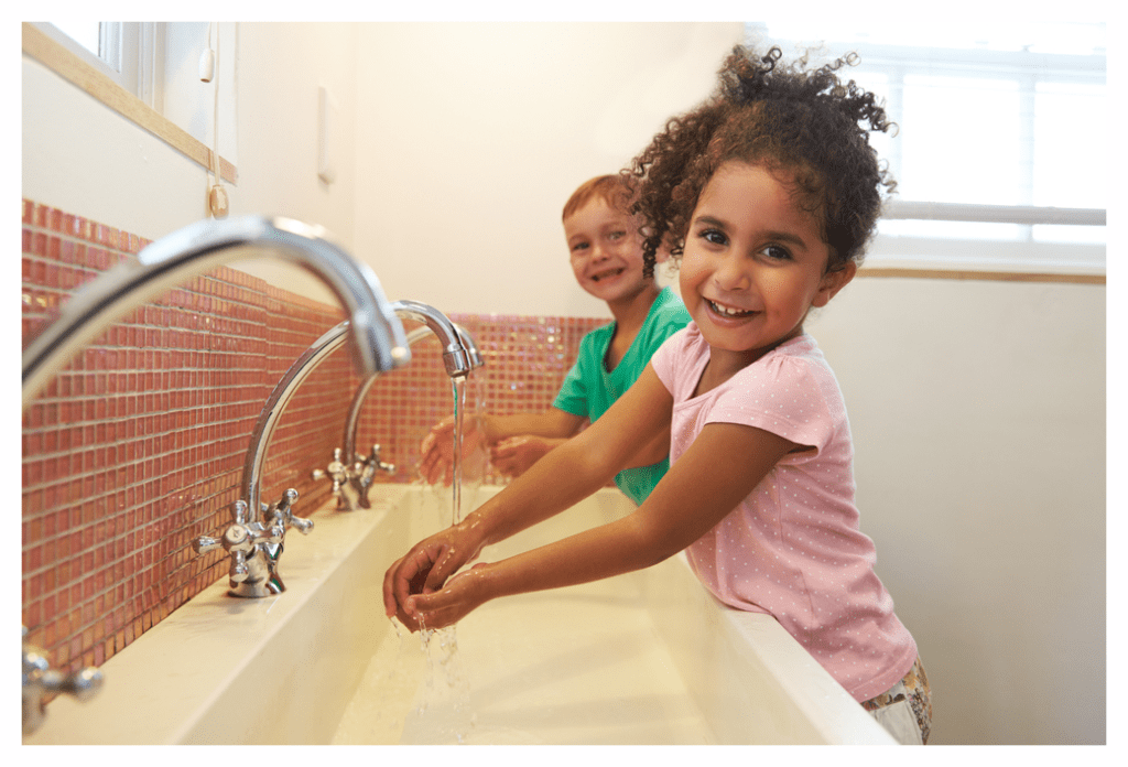 Two pre-school children washing their hands at a long sink with multiple taps. It's obviously a staged phot as strangely there is no soap in evidence.