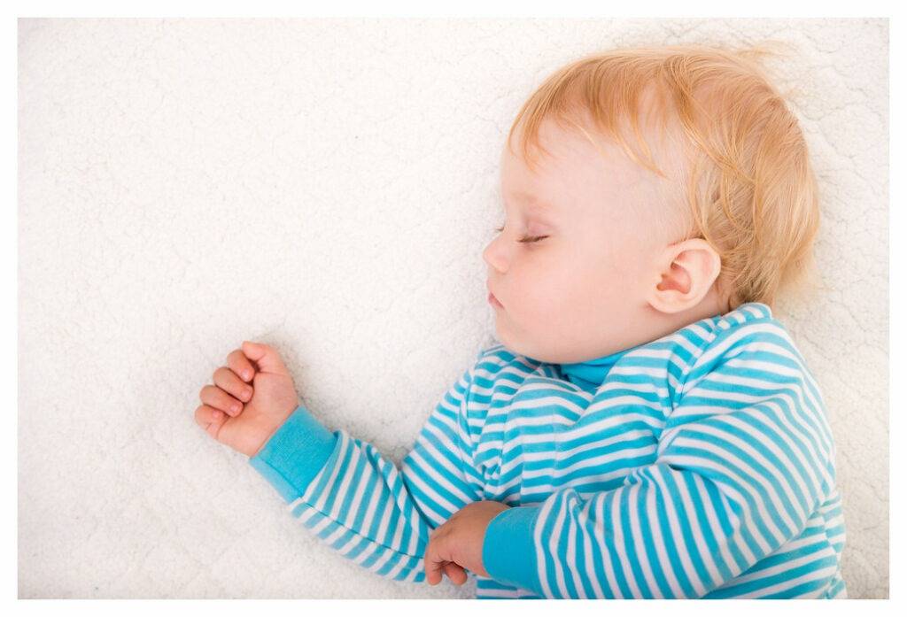 Young toddler in blue and white striped top sleeping on his back. He is sound asleep and looks like he is dreaming about boxing! 