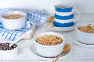 Eczema friendly pear oaty crumble desserts served in individual ceramic, white ramekins, with a side of carob nibs and classic blue and white striped jug of dairy free cream.