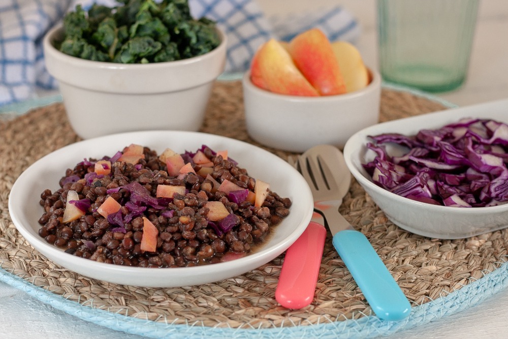 Puy lentils with red cabbage and apple served for a young child in a white bowl with brightly coloured children's cutlery.