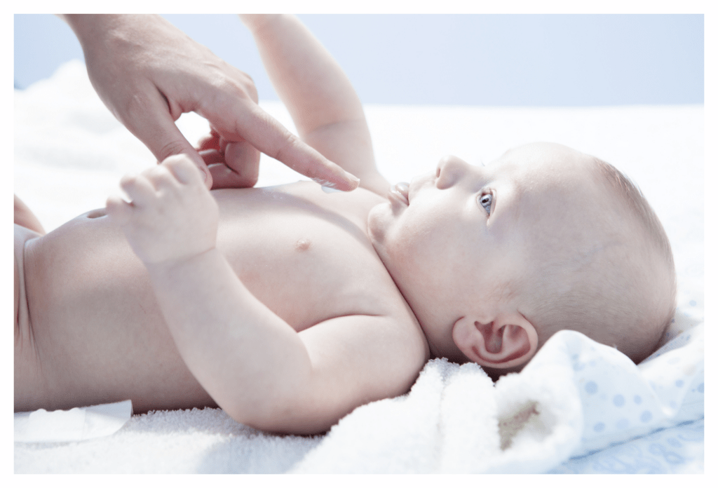 Adult applying 1 finger tip unit of topical steroid cream to chest of baby lying on white blanket and looking at adult trustingly